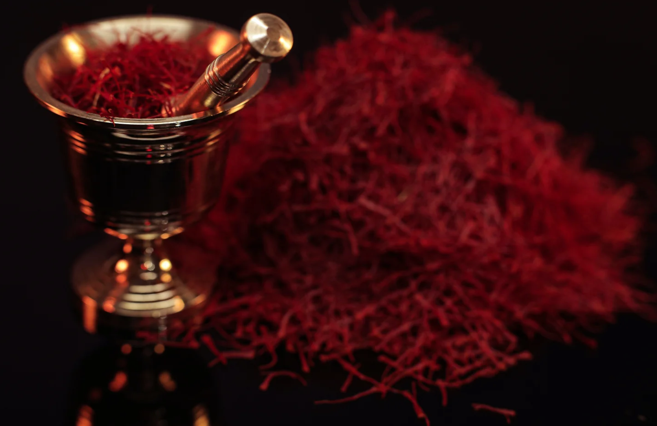 A pile of saffron strands on the right. A gold mortar and pestle with saffron inside it on the left.